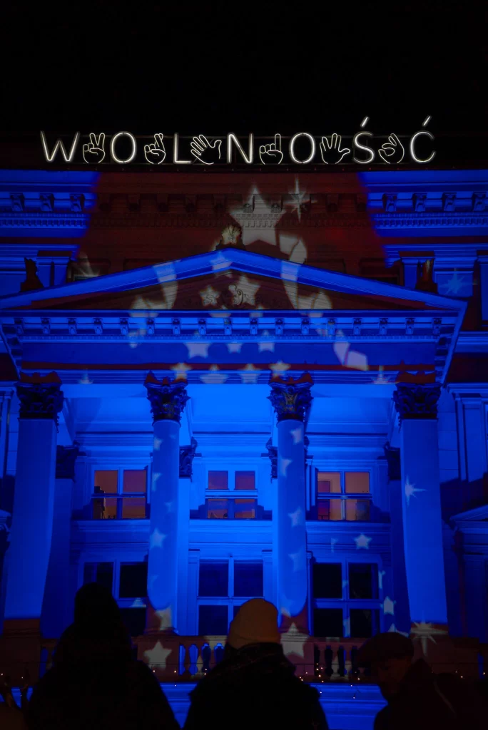 Front of a building that is illuminated in blue light with the words Wolnosc on the front. Heads of passerbys are shown 