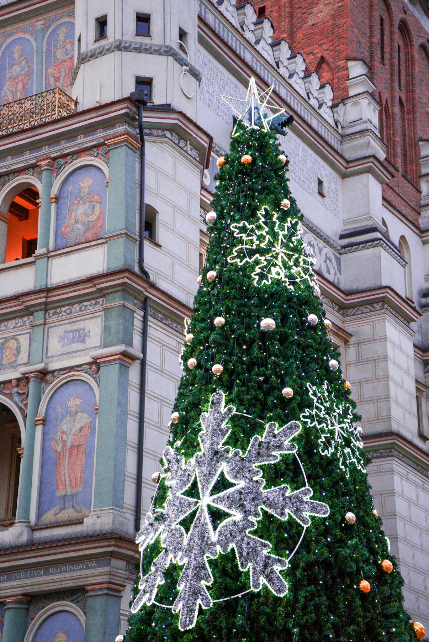 A lit up green christmas tree with silver snowflake lights attached to it set against the Poznan Town Hall or Ratusz