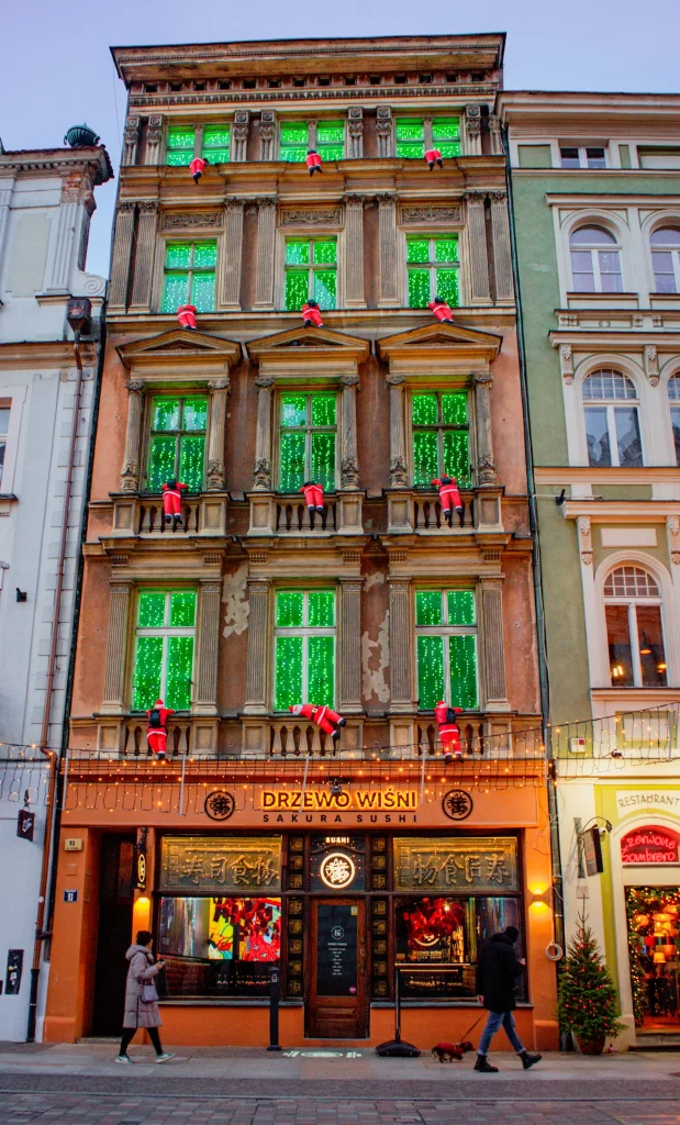Facade of a building in Poznan with miniature santas in redsuits hanging from the windows looking as if they are climbing into them. Windows are lit up in green