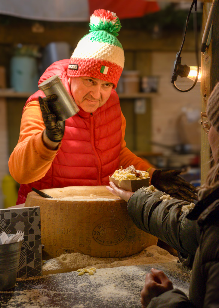 A Man in a Green, White and Red Beanie and red and orange sweater is serving cheese to patrons