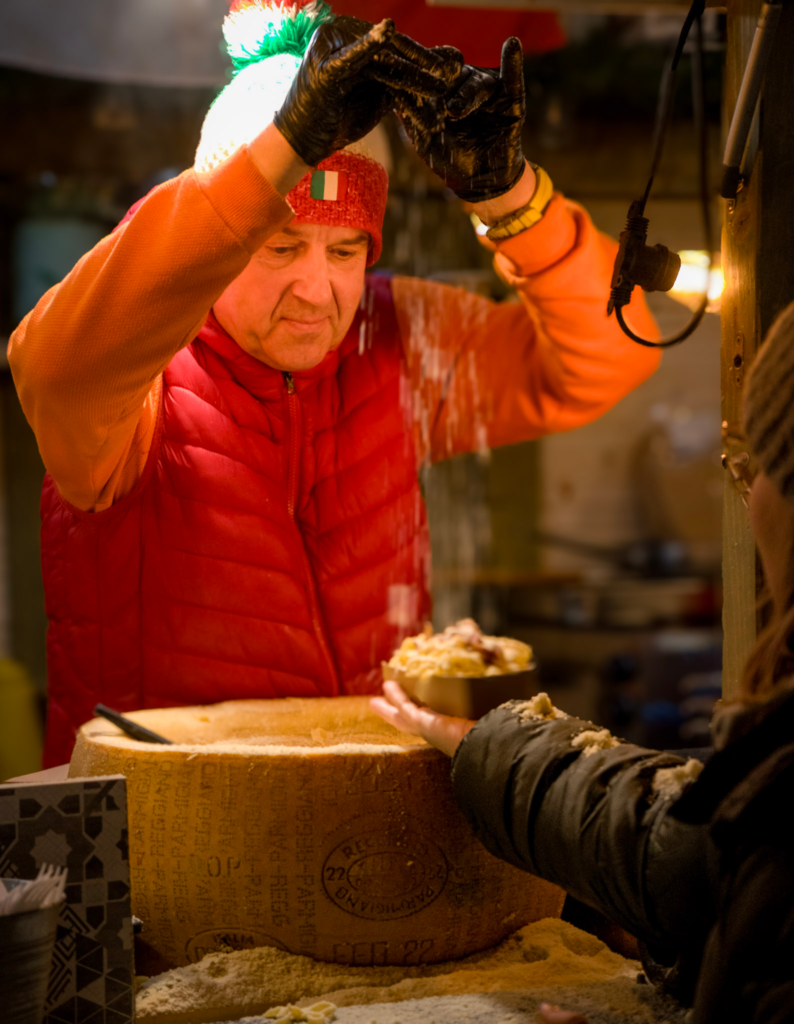 A Man in a Green, White and Red Beanie and red and orange sweater is serving cheese to patrons