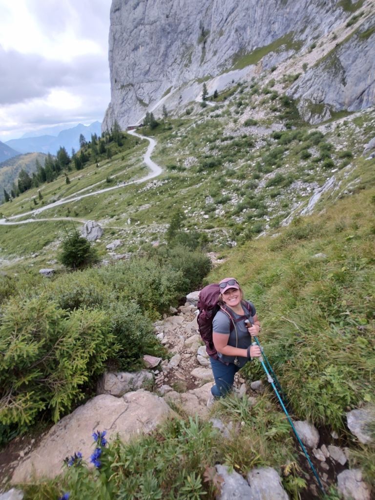 Woman in a pink hate with a grey shirt on and hiking poles in hand smiles at a camera on a ridgeline on the MADE trek in Italy