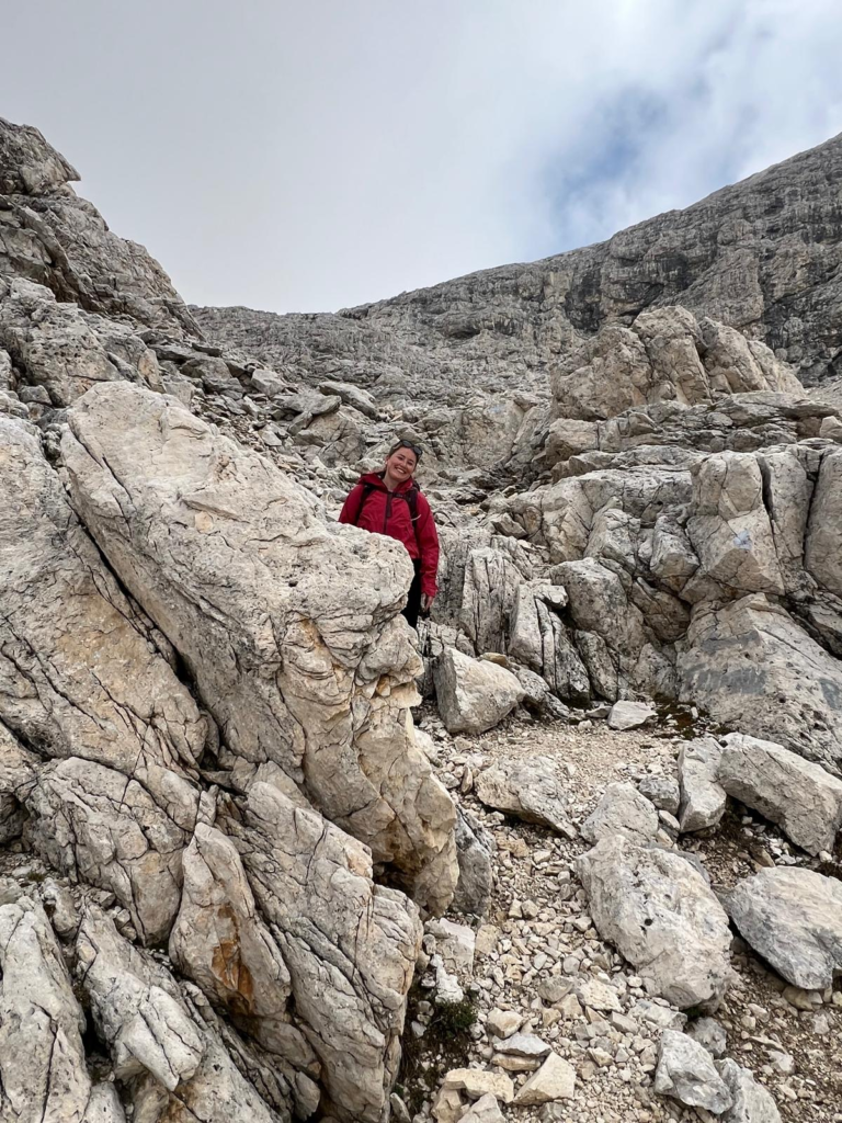 woman in red coat with her head playfully peeking from the rock and smiling at camera in a boulder field on Day 2 of the Palaronda Trek on the way to Rifugio Pradidali