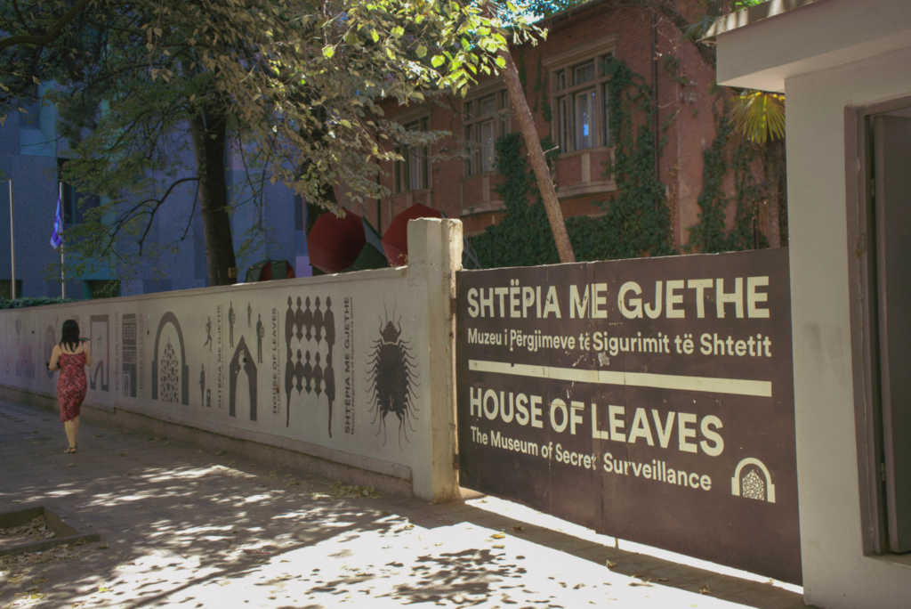 a building fence that says in black and white HOUSE OF LEAVES with trees in the background