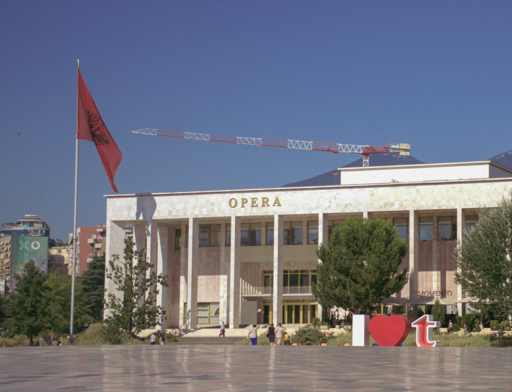 the opera house in Tirana Albania with giant albanian flag flying while tourists walk by
