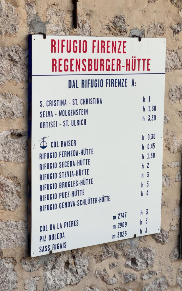 Multilingual signboard mounted on stone at Rifugio Firenze lists distances to nearby rifugi and peaks, including Rifugio Genova, Rifugio Stevia, and Sass Rigais.