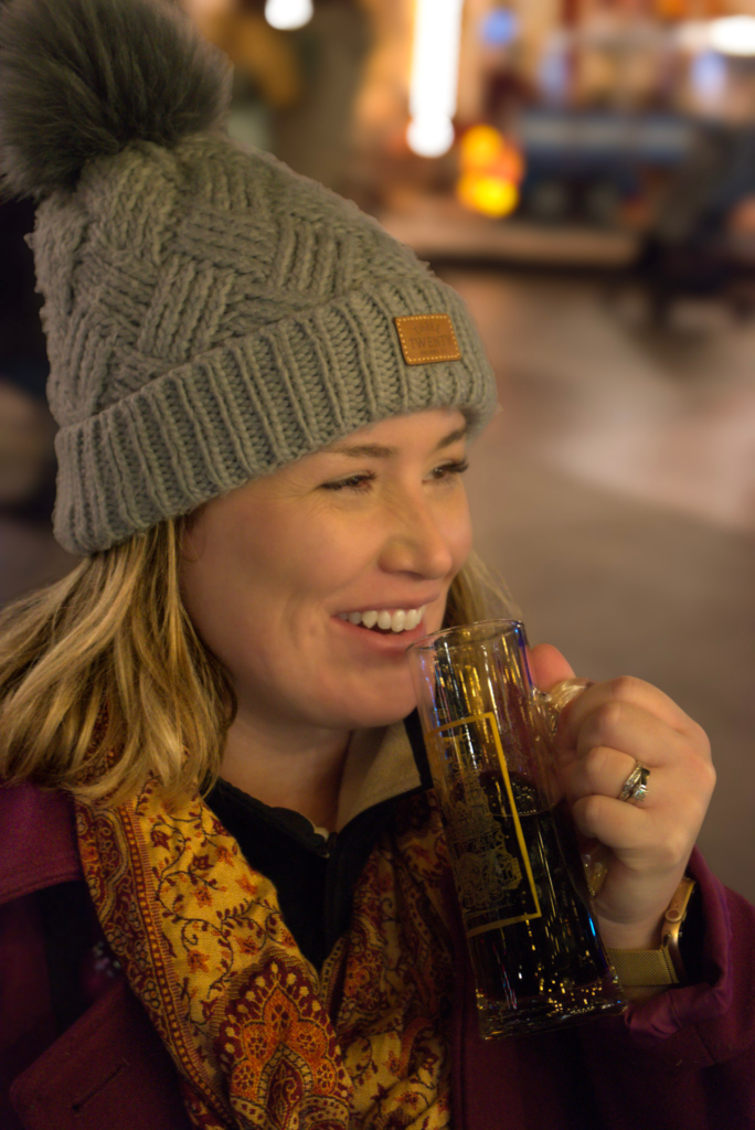 a woman with a grey beanie on, pink jacket and paisely colored scarf smiles as she drinks mulled wine in Bydgoszcz