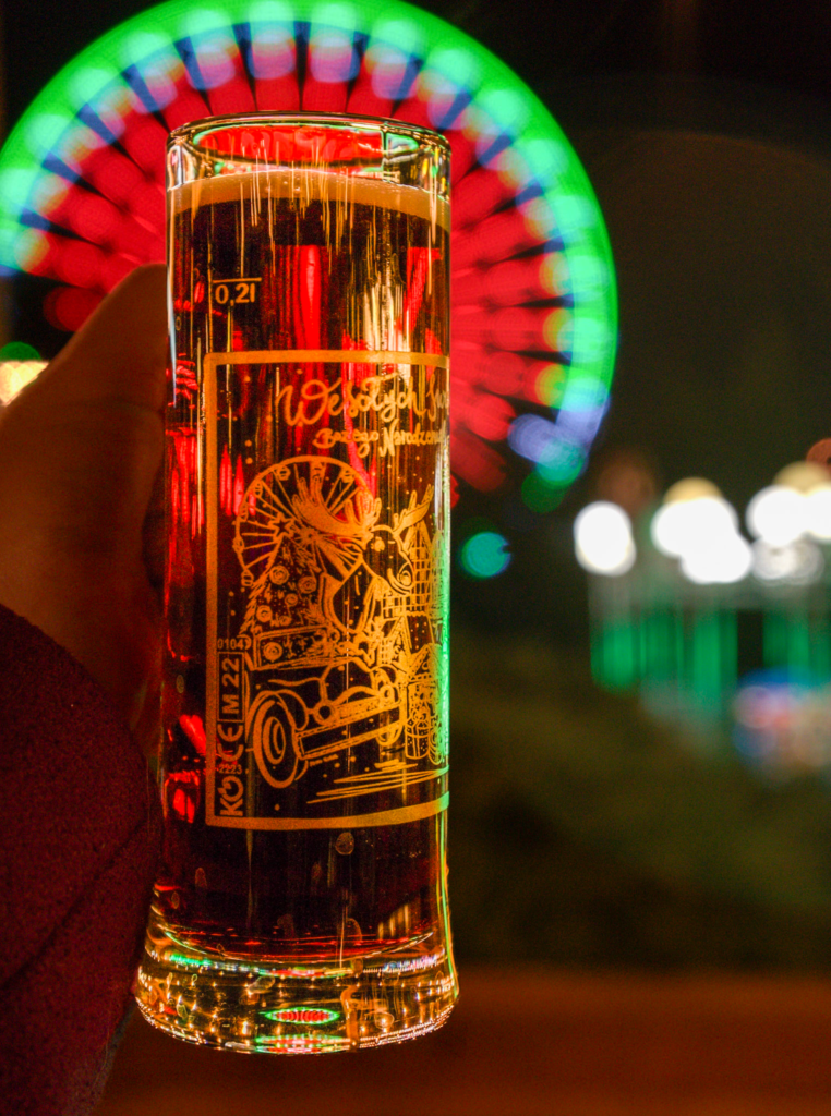 A tall christmas market mug with writing on it in Polish that says Bydgoszcz Christmas Market in Gold writing with a blurry Ferris wheel in the back