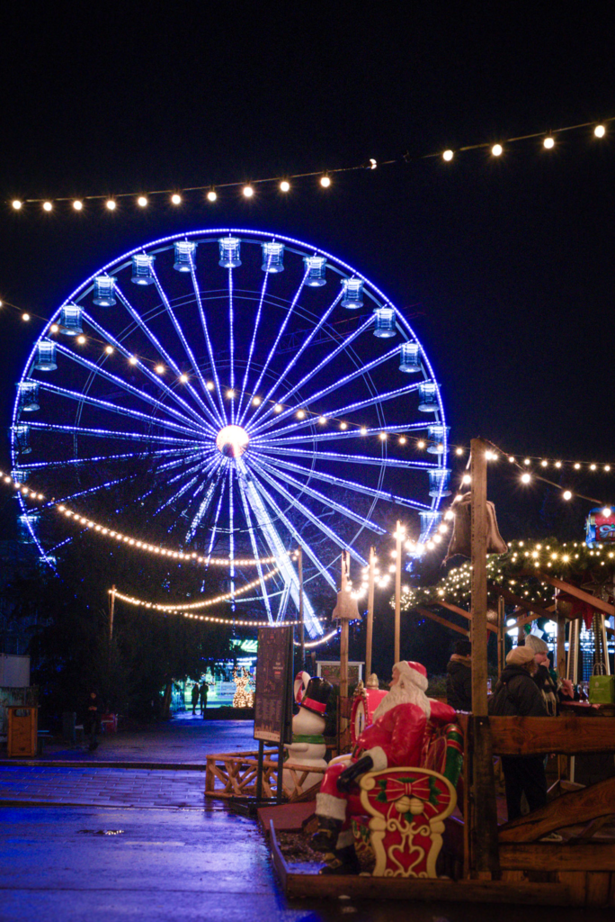 Ferris wheel lit up with blue lights while a plastic santa sits on a bench in the dark night at the Bydgoszcz Christmas Market