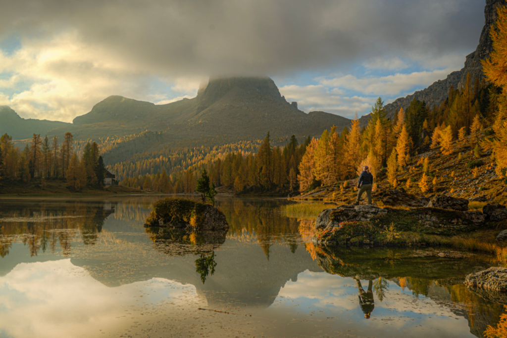 lago federa during the fall with bright orange hues bouncing off the lake early morning while a person stands on a rock watching the sunrise