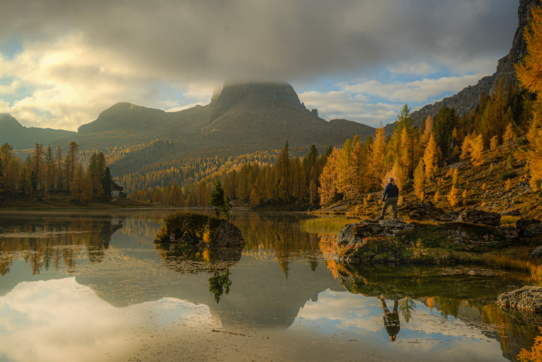 lago federa during the fall with bright orange hues bouncing off the lake early morning while a person stands on a rock watching the sunrise