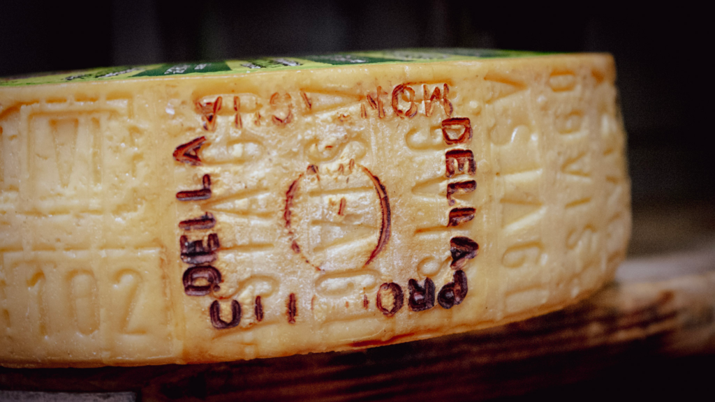 A close-up of a wheel of aged Parmigiano Reggiano cheese resting on a wooden shelf, showing its textured rind imprinted with certification markings and the words "PRODOTTO DELLA" and "ORIGINE." The yellow rind is dry and slightly cracked, highlighting the cheese’s authenticity and aging process.