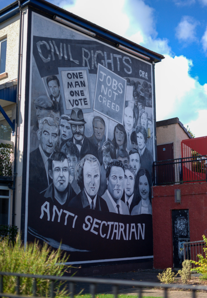 Large black and white mural on the side of a building in Derry Londonderry depicting civil rights leaders and community members beneath the words Civil Rights CRA and Anti Sectarian. Two protest signs in the artwork read One Man One Vote and Jobs Not Creed, referencing the Northern Ireland civil rights movement. The mural rises above a residential street with blue sky and neighboring buildings visible around it.