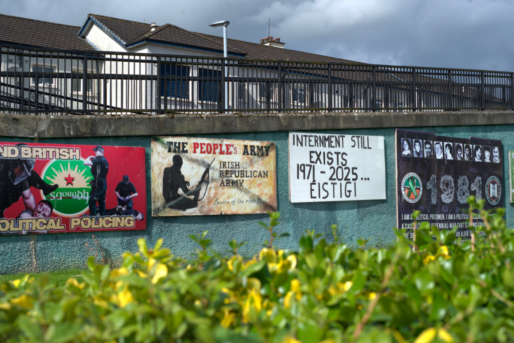 Murals in the Bogside neighborhood of Derry Londonderry Ireland line a concrete wall beneath a row of houses. The posters include text reading The Peoples Army Irish Republican Army and Internment Still Exists 1971 to 2025 Eistigi alongside images of armed figures and portraits referencing political prisoners. Green shrubs blur the foreground, grounding the scene in a residential area marked by historic and political imagery.