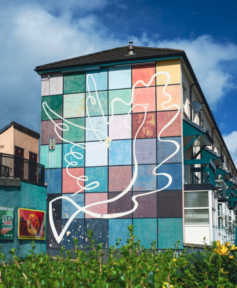 Colorful peace dove mural in the Bogside in Derry, painted as a white outline across a grid of teal, blue, pink, and green squares on the side of a residential building. The Bogside mural reflects themes of peace and reconciliation connected to the Bogside walking tour and the history of Bloody Sunday, making it a powerful stop on a one day Derry itinerary.