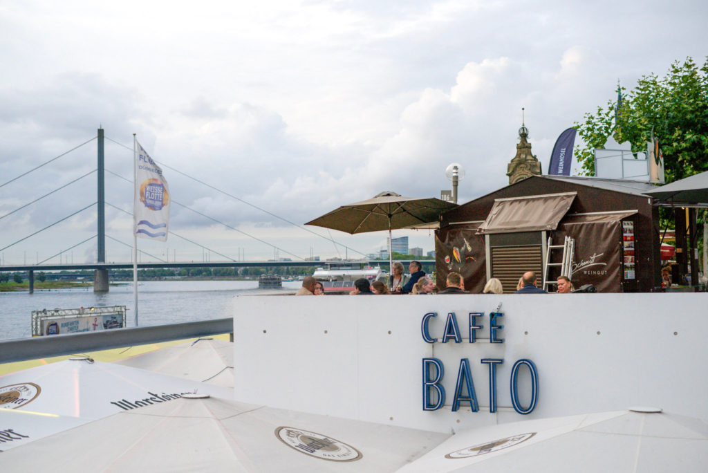 a white wall with a sign that says Cafe Bato in Dusseldorf Germany with people at the cafe indulging in drinks in the afternoon