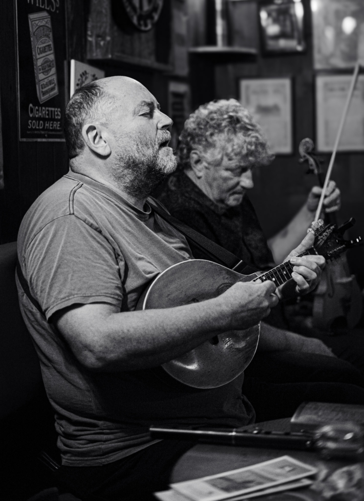 black and white photo of man singing with a small instrument and a man sitting next to him looking on at Olde Glen Pub in Donegal County, Ireland