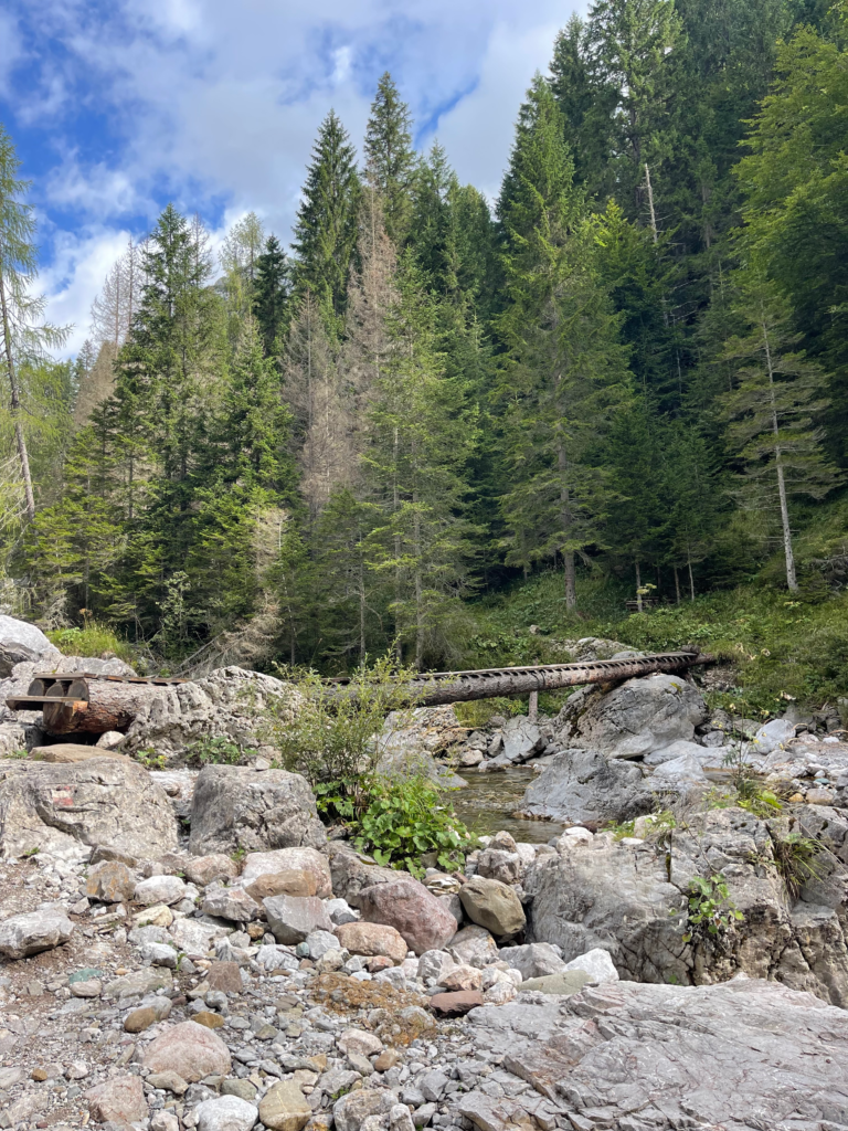 a wooden bridge over a river crossing with pine trees in the background and boulders strewn about