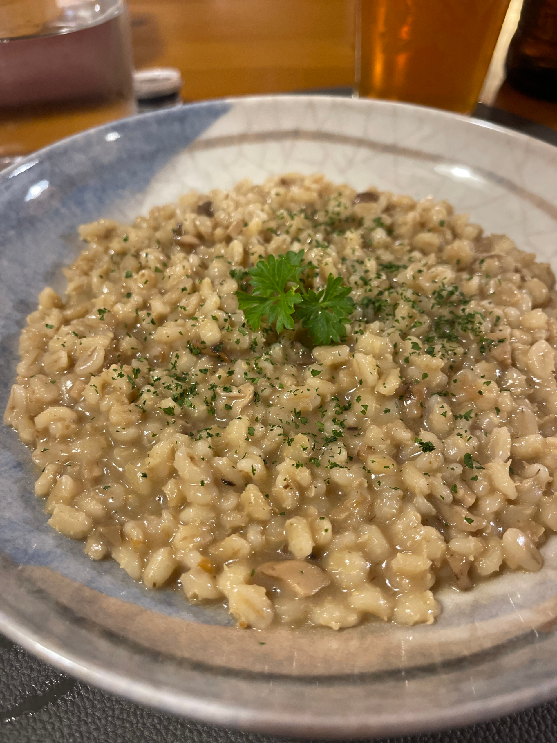 Creamy barley risotto with mushrooms served at Rifugio Tolazzi, garnished with fresh parsley. The dish is presented in a rustic bowl with a glass of amber beer in the background.