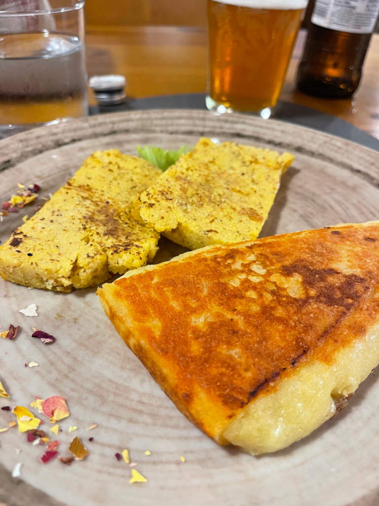 Frico served at Rifugio Tolazzi on the MADE trek, featuring a golden, crispy wedge of melted cheese alongside grilled polenta slices. The dish is garnished with dried flower petals, with a glass of beer and water in the background.