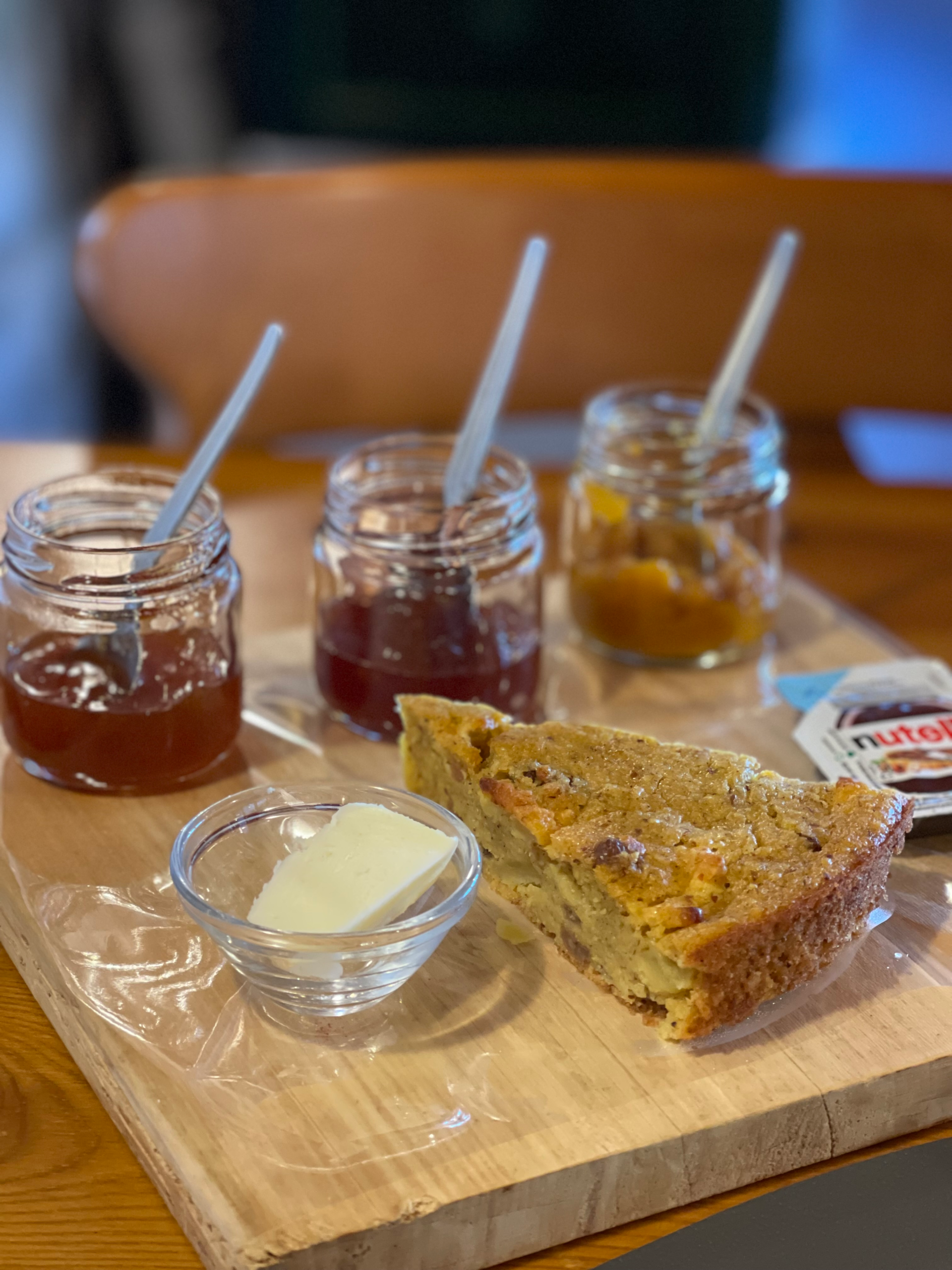 Slice of rustic polenta breakfast cake served on a wooden board, accompanied by a pat of butter, a Nutella packet, and small jars of assorted fruit jams with spoons. The cake appears dense and golden with visible bits of dried fruit or nuts.