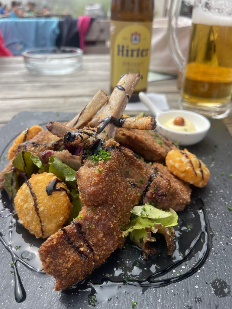 Breaded lamb chops served at Rifugio Marinelli, plated on a dark slate with crispy fried cheese balls, mixed greens, and a drizzle of balsamic glaze. A side of dipping sauce, a bottle of Hirter beer, and a glass of beer are visible in the background on a wooden outdoor table.
