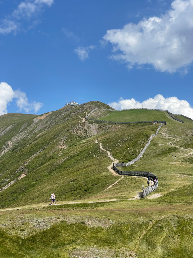 Hikers follow a winding alpine trail up a grassy ridge lined with wooden fencing on Alta Via 2, with a mountain hut and communications tower visible at the summit under a bright blue sky.