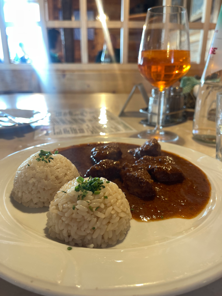 Plate of tender beef stew in rich brown sauce served with two scoops of herbed rice at a mountain hut along Alta Via 2, with a glass of Aperol Spritz in the background.