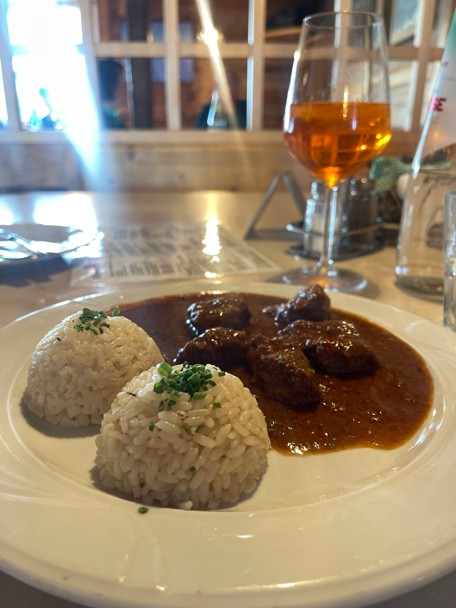 Plate of tender beef stew in rich brown sauce served with two scoops of herbed rice at a mountain hut along Alta Via 2, with a glass of Aperol Spritz in the background.