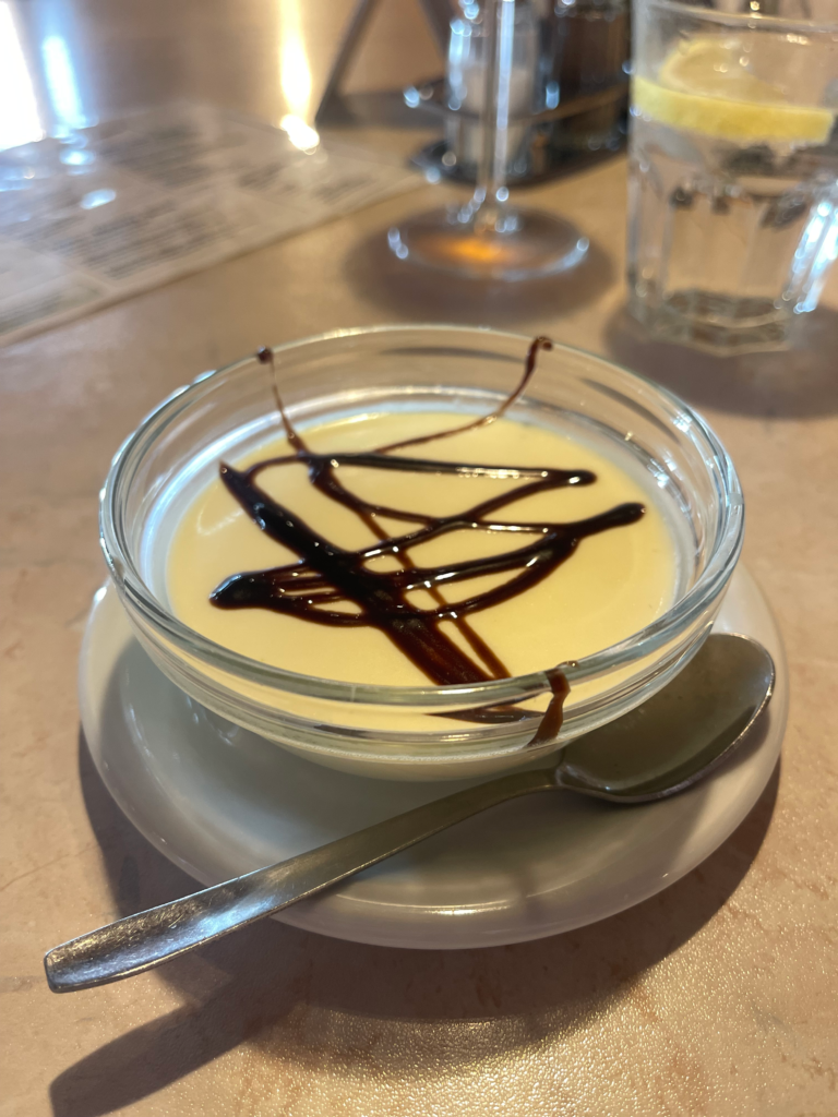 Glass bowl of creamy panna cotta drizzled with chocolate sauce served on a white saucer, photographed inside a rustic rifugio on the Alta Via 2 trail.