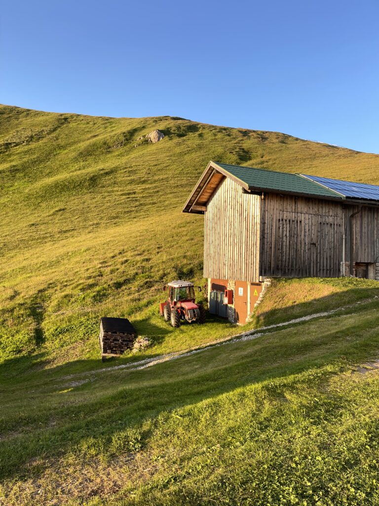 Barn and tractor near Rifugio Genova: Red tractor parked beside a wooden alpine barn with a green roof, surrounded by sunlit pastures near Rifugio Schlüterhütte (Rifugio Genova) on Day 2 of the Alta Via 2 trail.