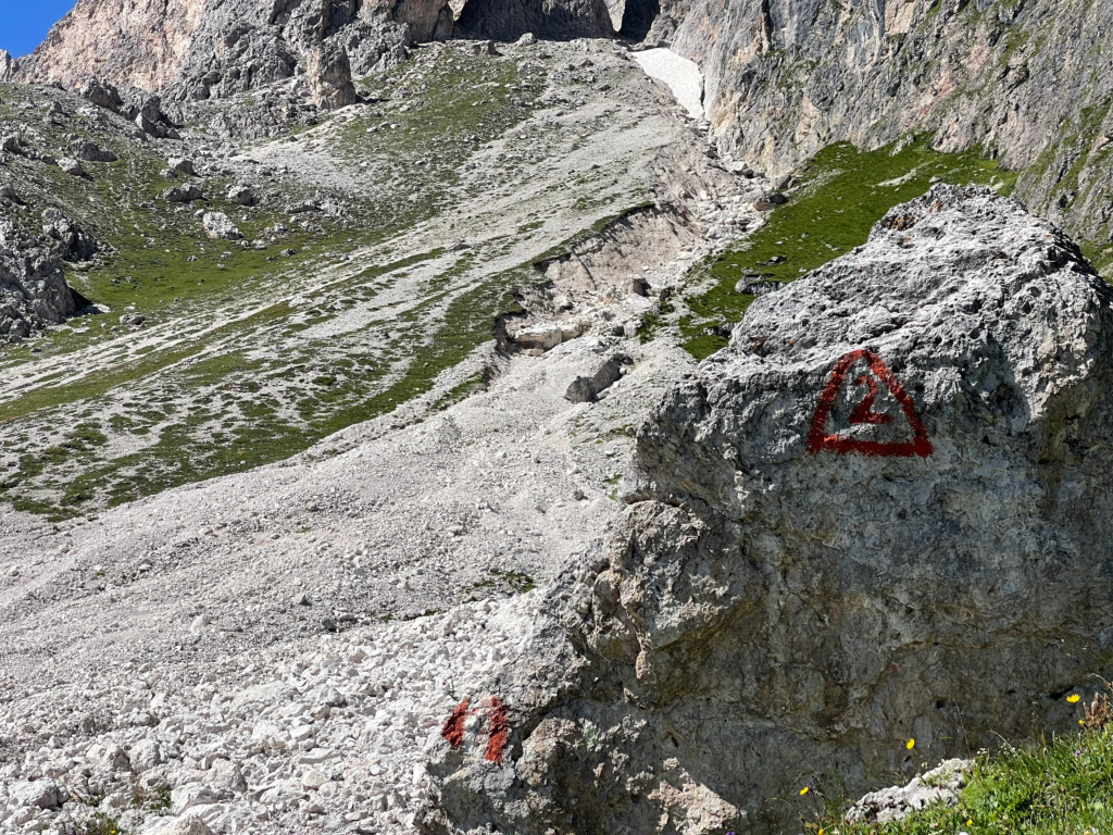 Painted red triangle with the number 2 marks the Alta Via 2 trail on a boulder near a steep, rocky ascent to Forcella Roa pass between Rifugio Genova and Rifugio Firenze.