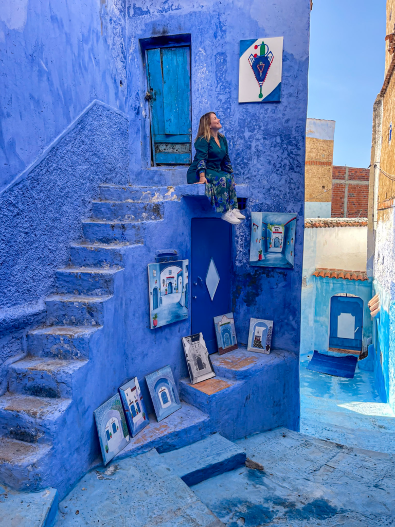 woman in a blue alleyway of chefchaouen morocco sitting on a ledge surrounded by paintings