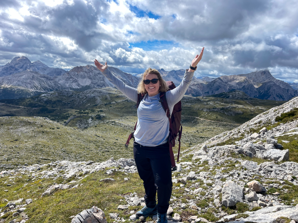 Woman in grey longsleeved shirt with hiking bag and black pants has arms outstretched infront of Rifugio Seekofel