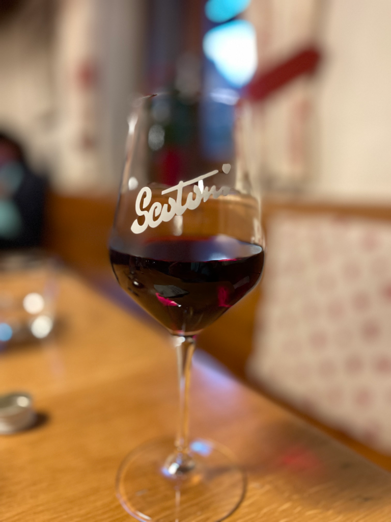 Glass of red wine served at Rifugio Scotoni, in a tall stemmed glass branded with “Scotoni” in white script. The glass rests on a wooden table in a warm, rustic indoor setting with soft, ambient lighting.