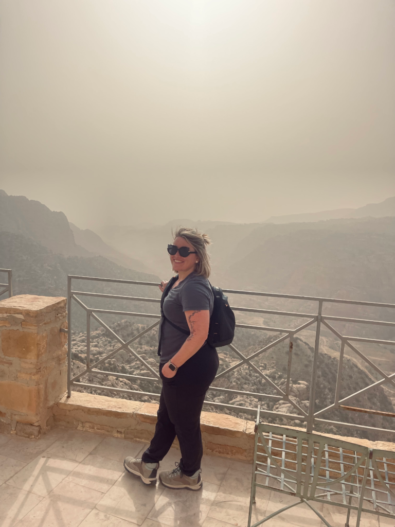 Woman in a dark shirt and pants smiling at camera standing near railing at the Dana Biosphere in Jordan with a hazy view in the background.