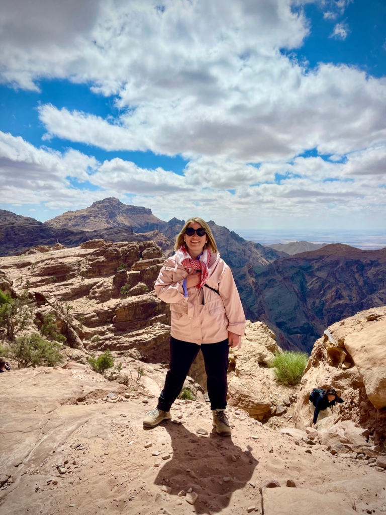 woman with a pink jacket on and black pants stands on the edge of a canyon in Petra during the petra back trail hike