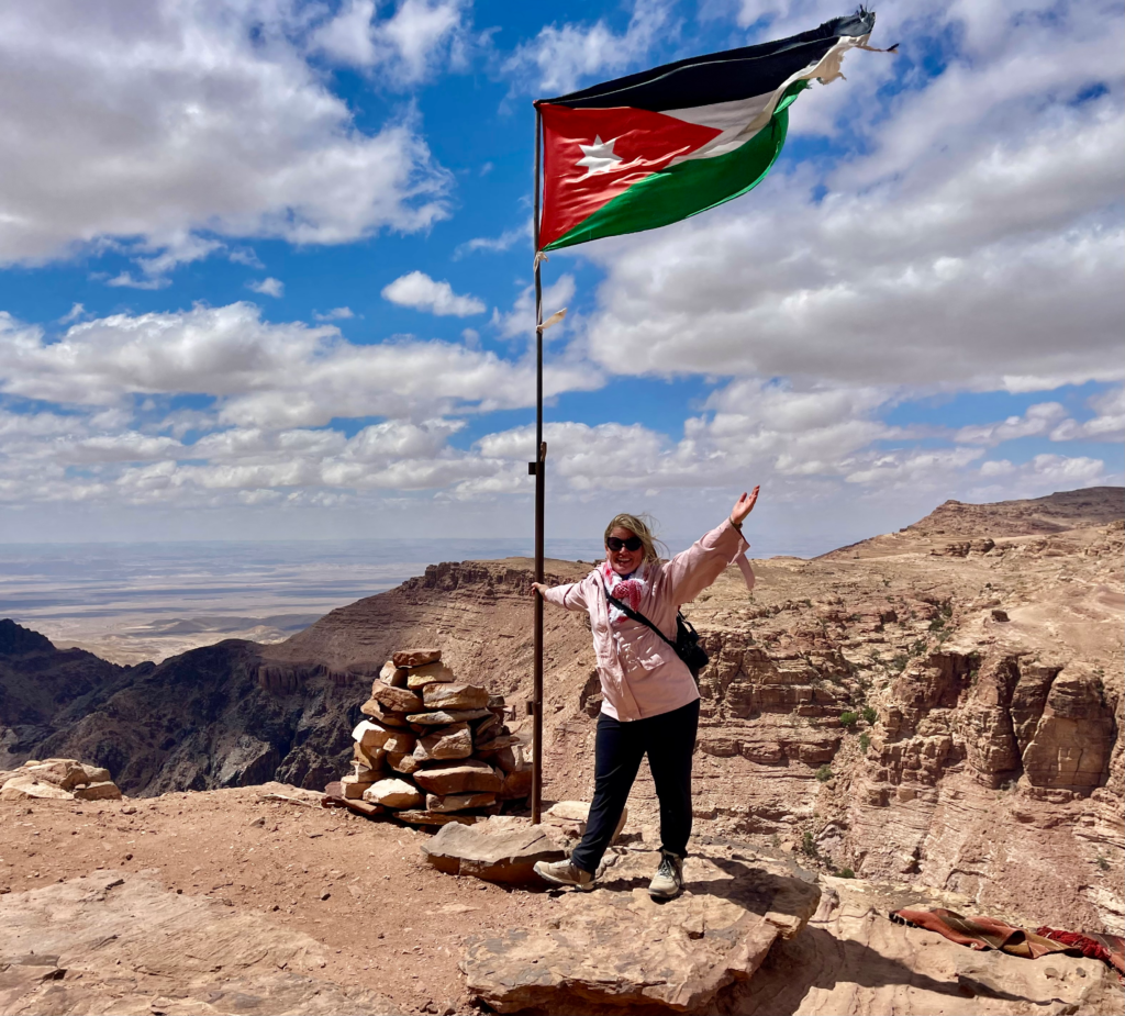Woman with pink jacket and black pants on holds on to a flag pole in Petra with the Jordanian Flag