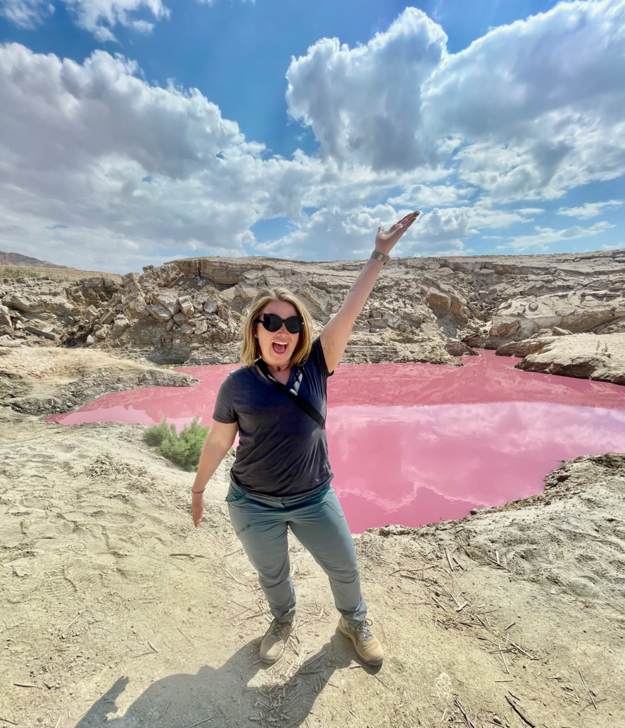 woman with outstretched arms in hiking gear in front of pink pools of the Dead Sea