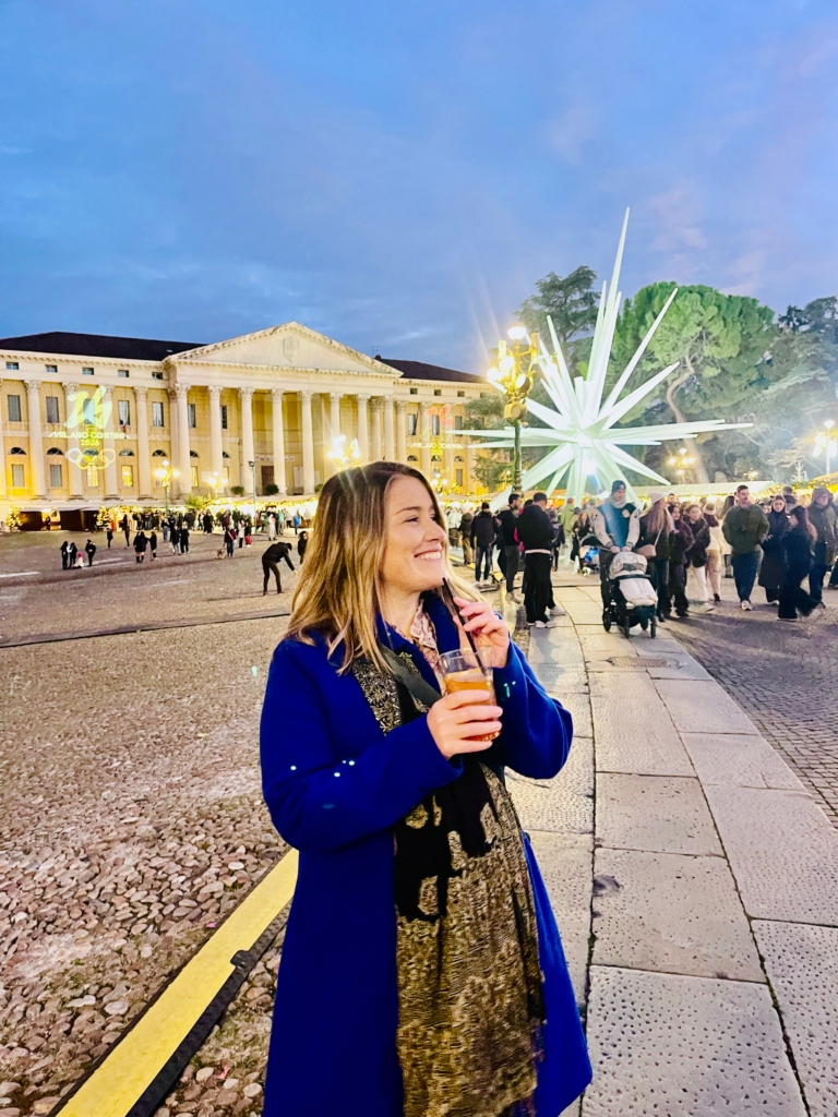 woman in a blue coat and black and gold scarf with blonde hair standing in front of a christmas market stall in Verona Italy with an Aperol Spritz in her hand