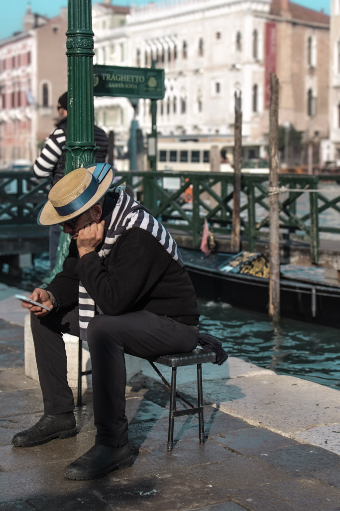 A Gondolier sits looking down at his phone in his tradition white and red top with venetian hat as he waits for his next customer near the Traghetto