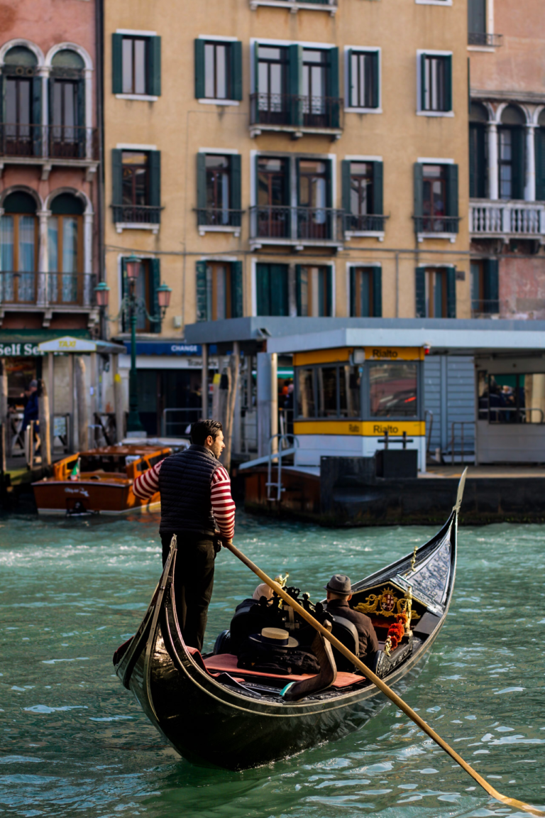 A Venetian Gondolier rows his gondola in the waters of venice as two people sit in his boat