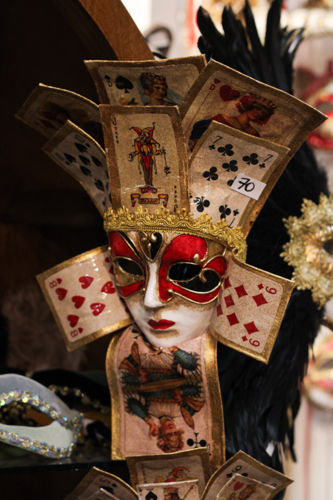 a colorful mask in red and white with gold cards and playing cards surrounding it hanging on a wall