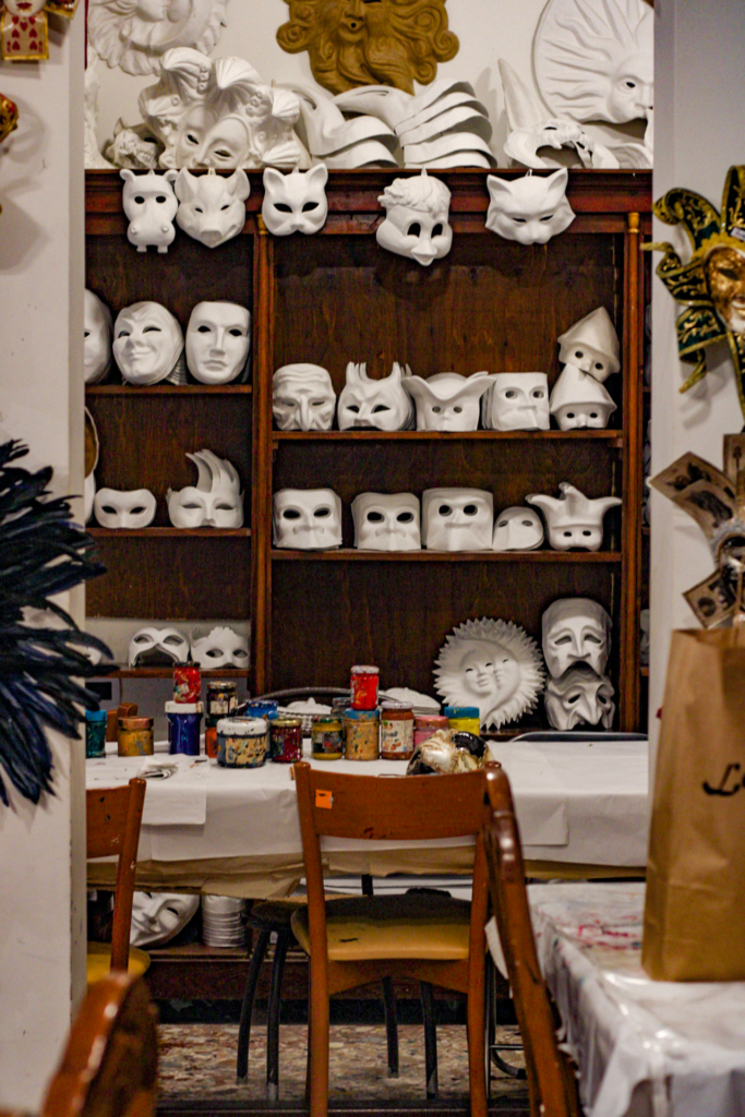 a mask making workshop with a table for decorating masks behind the table is a selection of white paper mache masks one can choose for their mask making tour in Venice