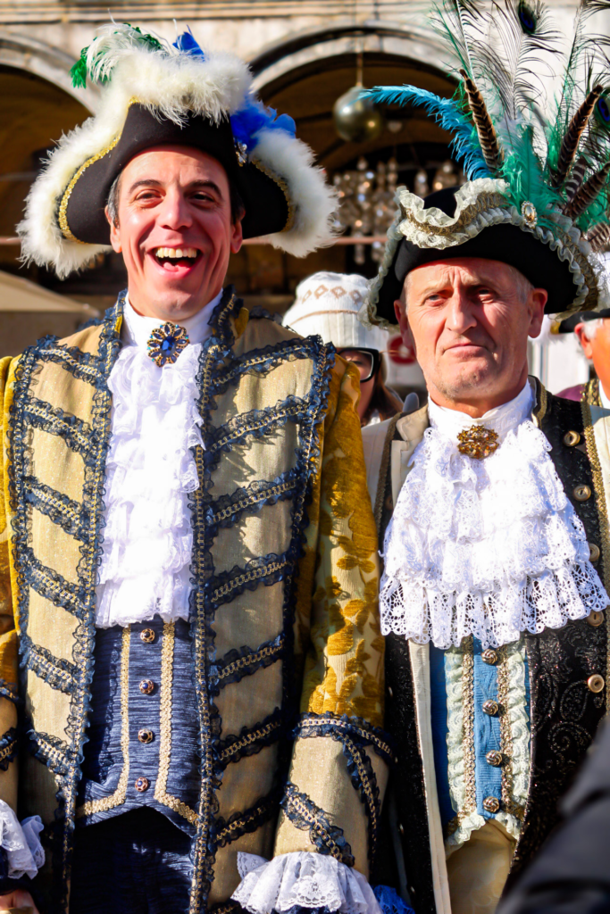 Venice Carnivale and two men smiling at the camera in st. marks square while wearing costumes with gold and black detail and 17th century style hats.