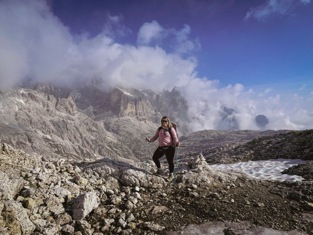 Woman in a pink shirt and wearing hiking gear holding trekking poles in her hand is posing against the Pala Group in the Dolomites