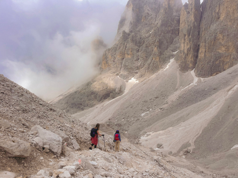 two hikers in dark clothing and backpacks head down a gravel path on a mountain that is surrounded by fog and scree fields on the 1st day of the Palaronda Trek in the Pale Di San Martino, Dolomites.