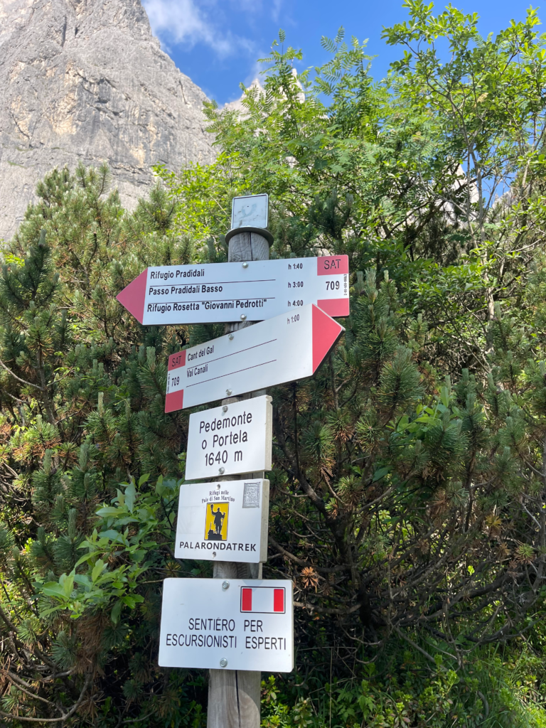 a trail market in white and red with times and distances in kilometers showing how far rifugio treviso, rifugio pradidali are with a Palaronda Trek sign beneath everything