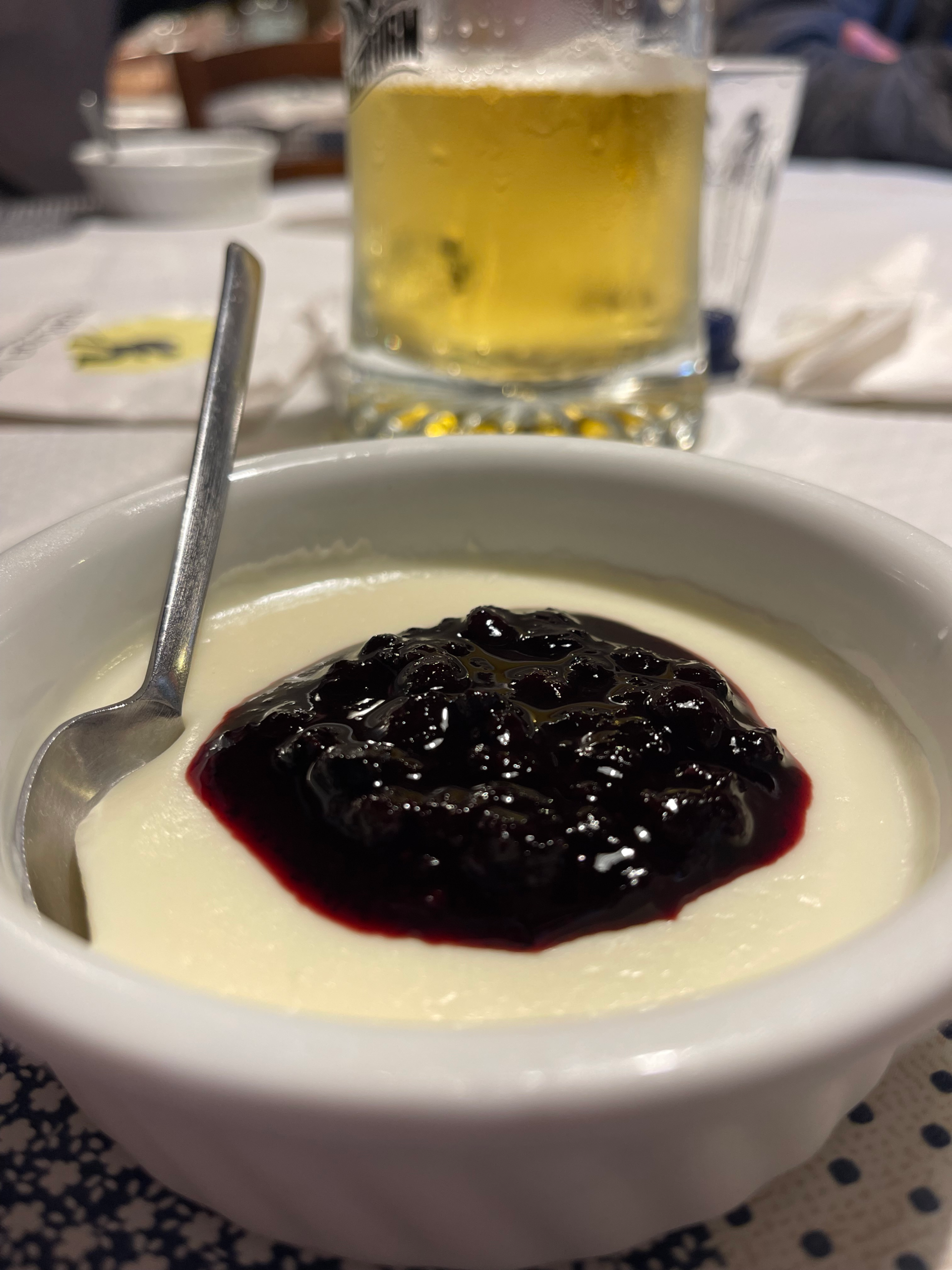 Creamy panna cotta served at Rifugio Treviso, topped with a generous layer of dark berry compote. A spoon rests in the dish, with a cold glass of beer in the background on a white tablecloth.