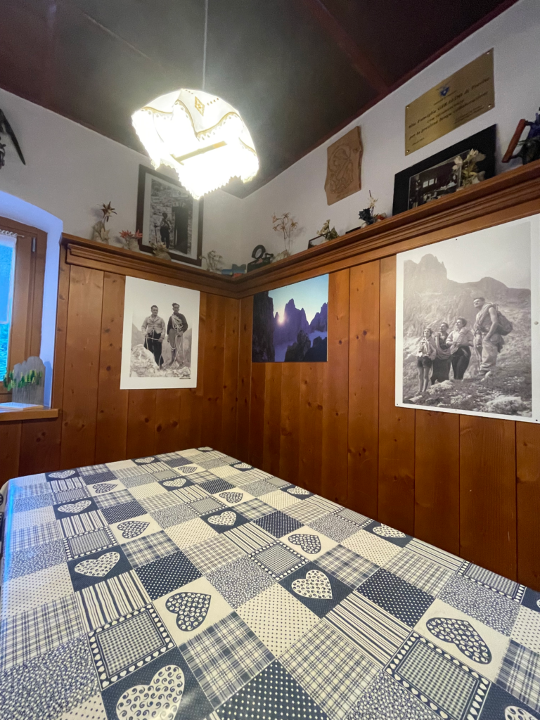 Cozy dining corner at Rifugio Treviso featuring a wood-paneled interior, a patterned blue-and-white tablecloth with heart and checkered designs, and vintage black-and-white mountain climbing photos on the wall. A warm overhead light adds to the rustic alpine charm.